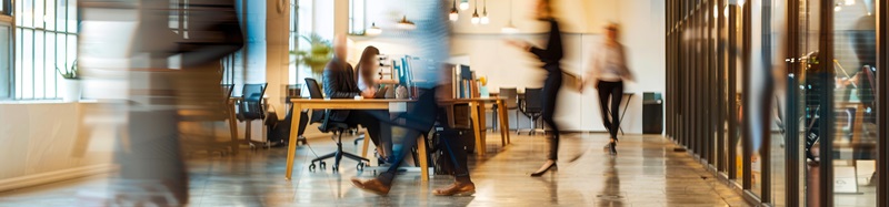 This photo captures a modern office space with several blurred figures of business professionals going about their work.